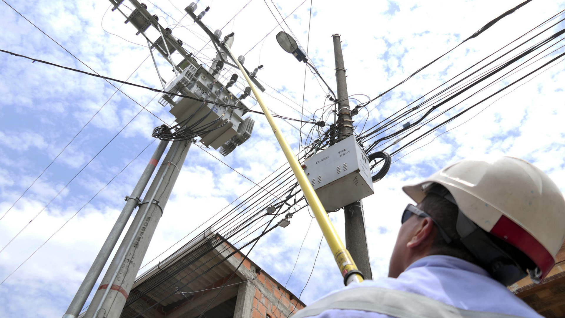 Hombre con casco y camisa blanca de pie junto a un poste de electricidad, observando el entorno con atención.